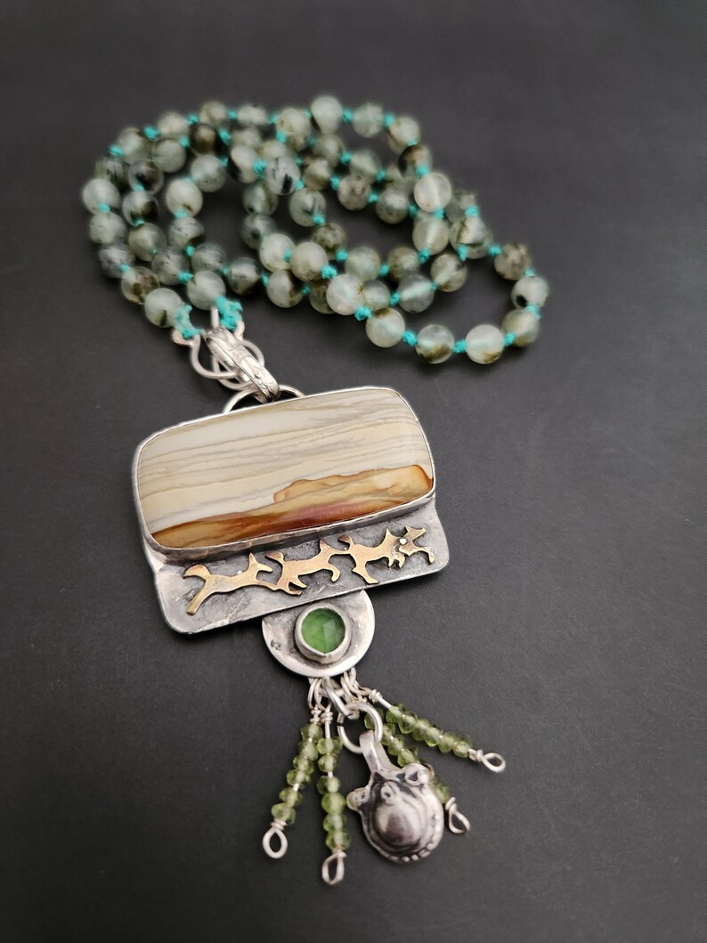May include: A silver and gold necklace with a large, rectangular, brown and white agate pendant. The pendant is surrounded by a silver frame with gold accents. The necklace is made of green beads and has a silver and gold charm with a green stone.