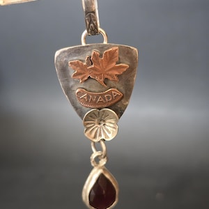 Puede incluir: Colgante de plata con un diseño de hoja de arce de cobre y la palabra "CANADA". El colgante tiene una gema roja oscura en forma de lágrima y un pequeño dije de flor. Cuelga de una argolla de plata.