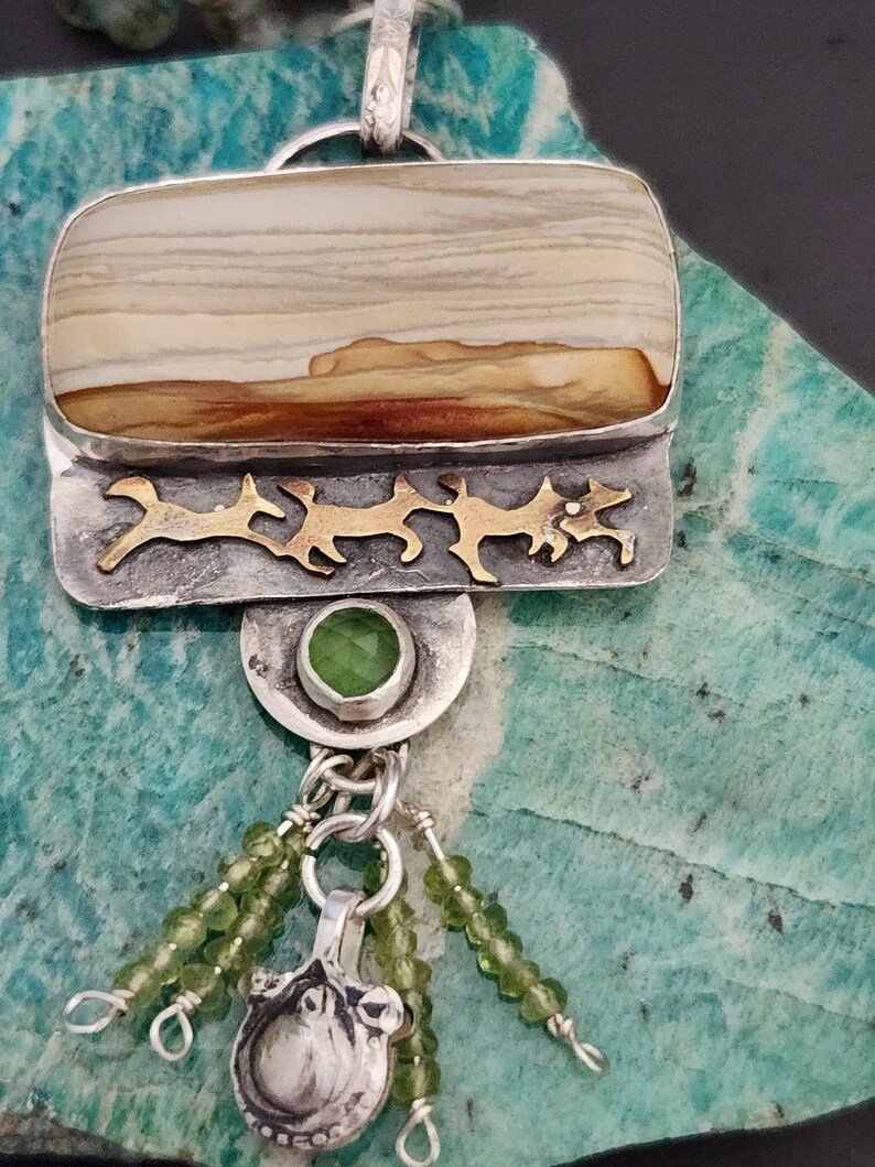 May include: A silver and gold pendant necklace with a rectangular agate stone, a green gemstone, and a silver charm. The pendant is hanging from a silver chain.