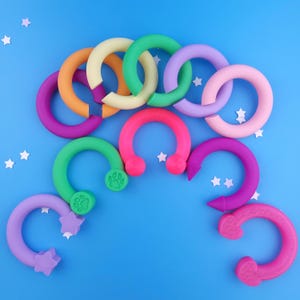 May include: A collection of ten C-shaped silicone rings in various colours including orange, cream, green, purple, pink, and magenta. Some rings feature decorative ends shaped like stars, paw prints, or pointed tips, arranged on a blue background.