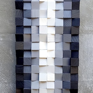 Könnte beinhalten: Ein Wandkunstwerk aus Holzblöcken in Grautönen, Weiß und Schwarz. Die Blöcke sind in einem Farbverlauf angeordnet und erzeugen ein visuell ansprechendes abstraktes Design.