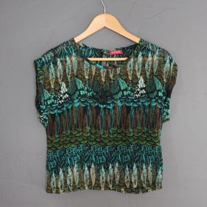 May include: A short-sleeved top with a colorful feather pattern in shades of teal, brown, and green. The top is hanging on a wooden hanger against a gray background. The top has a round neckline.