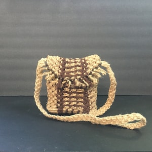 May include: A small, hand-woven crossbody bag in natural and brown tones. The bag features a rectangular shape with a flap closure and a long, adjustable strap. The design incorporates a striped pattern and fringe detailing, creating a rustic, bohemian aesthetic.