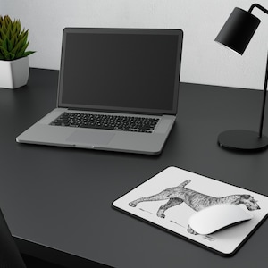 Op de afbeelding: Een rechthoekige witte muismat met een gedetailleerde zwarte inktillustratie van een hond in een staande profielpositie. De muismat heeft een dunne zwarte rand en ligt op een donker oppervlak naast een laptop en een witte computermuis.