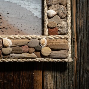 May include: A close-up of a picture frame made of driftwood, rope, and seashells. The frame is brown and beige and has a rustic look. The frame is set against a wooden background.