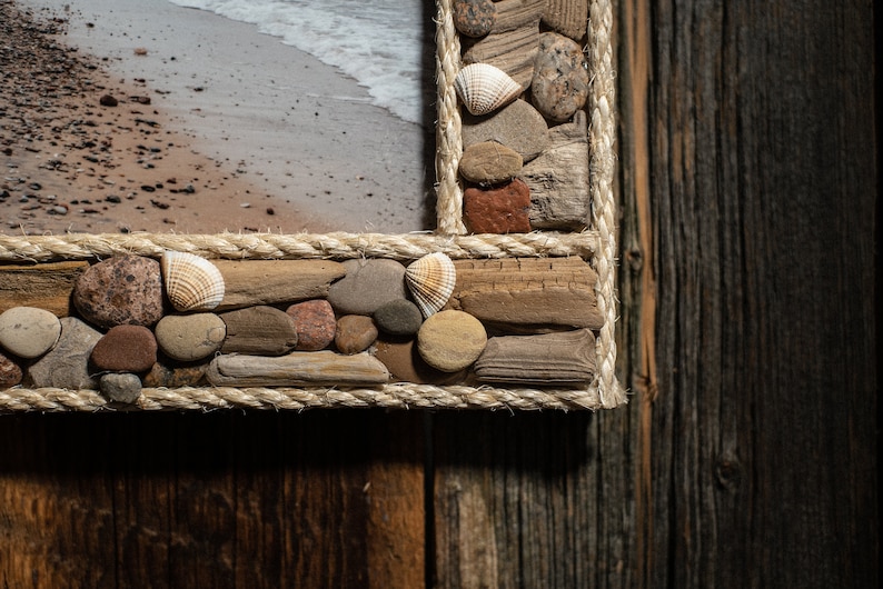 May include: A close-up of a picture frame made of driftwood, rope, and seashells. The frame is brown and beige and has a rustic look. The frame is set against a wooden background.