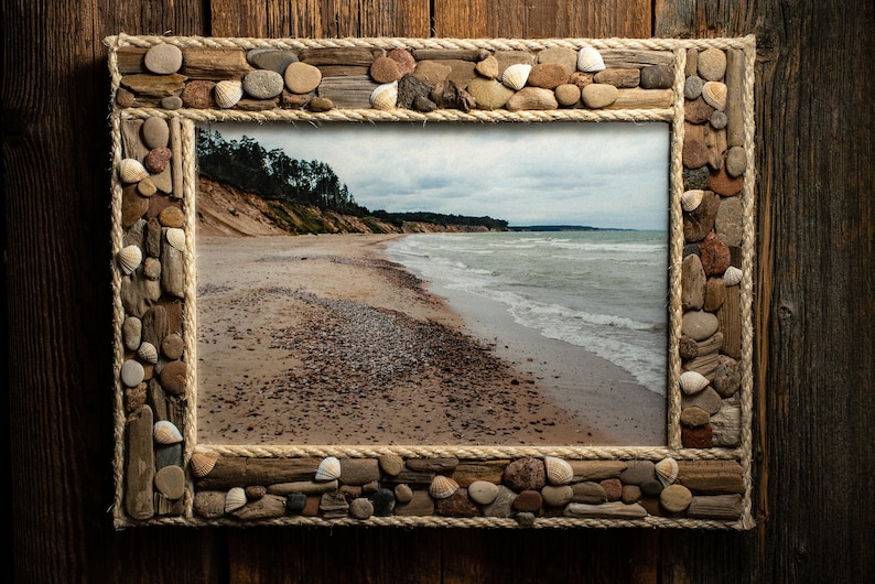 May include: A rustic wooden picture frame with a rope border and a seashell and stone design. The frame holds a photograph of a sandy beach with a blue ocean and a cloudy sky.