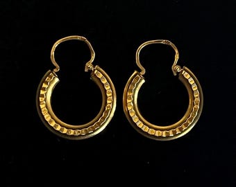 May include: A pair of gold hoop earrings featuring a textured, notched outer edge and a smooth inner band. The earrings are circular in shape and include a hinged wire closure mechanism at the top for securing the hoops in place.