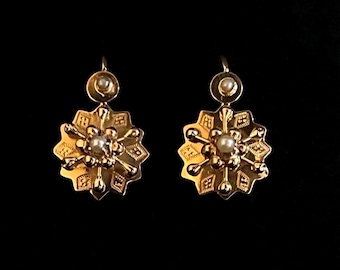May include: A pair of gold drop earrings featuring a star-shaped design with textured geometric patterns. Each earring has a central white pearl, radiating metal spokes, and a smaller circular top component set with a single white pearl.