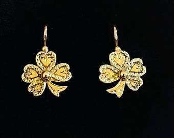 May include: A pair of gold-colored earrings. Each earring features a clover-shaped design with a small, round center and a bow-like detail at the bottom. The earrings are suspended from a simple hook.