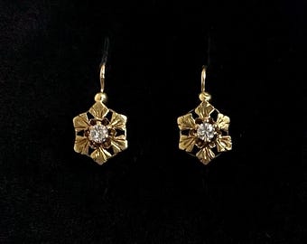 May include: A pair of gold drop earrings featuring a six-petaled floral or snowflake-shaped design. Each earring is centered with a single clear, round-cut gemstone and is suspended from a simple gold hook wire, creating a delicate jewelry piece.