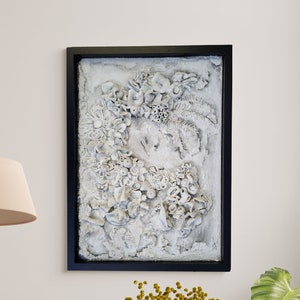 Può includere: Un'opera d'arte da parete incorniciata in bianco con un design testurizzato 3D di fiori e foglie bianche. L'opera d'arte è in una cornice nera.