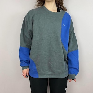 May include: A grey and blue Nike sweatshirt. The sweatshirt features a grey body with blue panels on the sleeves and side. A small white Nike swoosh logo is on the chest. The sweatshirt is a crew neck style.