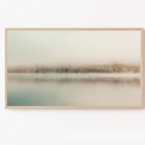 Puede incluir: Una fotografía enmarcada de un lago brumoso con una línea de árboles en el fondo. El agua es de color azul claro y el cielo es de un blanco suave.