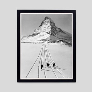 May include: Black and white photograph of a snowy mountain peak with three skiers in the foreground. The skiers are walking up a path in the snow, leaving tracks behind them.