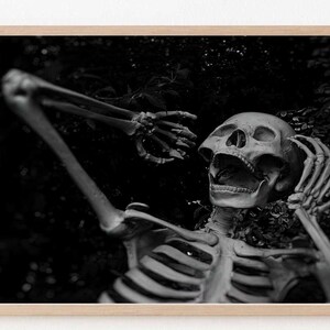 May include: A black and white photograph of a human skeleton lying on its back with its mouth open in a scream. The skeleton is surrounded by dark foliage.