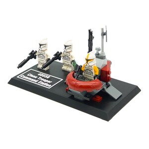 Op de afbeelding: LEGO Star Wars 40558 Clone Trooper Command Station set met drie witte clone trooper minifiguren, één in een rode en grijze speeder, en een zwarte basis met de tekst "40558 Clone Trooper Command Station".