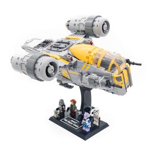 May include: A LEGO model of The Razor Crest starship, with grey, yellow, and white parts. The model includes several minifigures on a black base, with the text "75447 The Razor Crest".