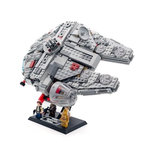 Puede incluir: Un modelo detallado de la nave espacial Halcón Milenario, construido con bloques de construcción grises y rojos. El modelo incluye figuras en miniatura de Han Solo, Chewbacca y C-3PO, expuestas en un soporte negro. Un objeto de colección de Star Wars.