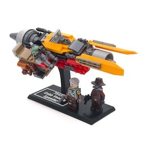 May include: A Lego model of Cobb Vanth's Speeder, with yellow, grey, and red parts. Two Lego minifigures are positioned in front of the speeder on a black base. The base has text that reads "75312 Cobb Vanth Speeder".