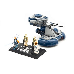 Può includere: Un set Lego Star Wars con un carro armato d'assalto corazzato blu e grigio con tre minifigure. Il set include un clone trooper, un pilota clone e un comandante clone. Il set è esposto su una base nera con il testo "75283 Armored Assault Tank".