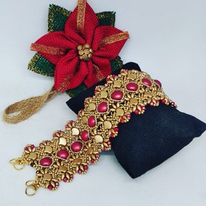 Può includere: Un braccialetto di perline dorate e bordeaux con un design decorativo. Il braccialetto è su una superficie di velluto nero.