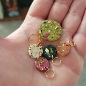 May include: Five colorful stitch markers with gold rings. The markers are round and have a speckled pattern in shades of green, white, pink, and brown.
