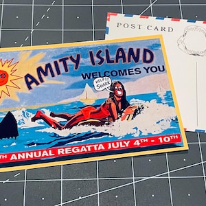 Peut inclure: Une carte postale rectangulaire avec une illustration colorée d'une personne faisant du surf, portant le texte AMITY ISLAND WELCOMES YOU, HELP SHARK et 50TH ANNUAL REGATTA JULY 4TH - 10TH. Le verso affiche POST CARD et un dessin de mâchoire de requin.