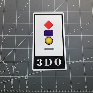 May include: A white sticker with a black border and the text "3DO" in white letters. The sticker features a red diamond, a purple square, and a yellow circle stacked on top of each other.