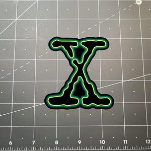 Könnte beinhalten: Ein schwarzer Aufnäher mit einer grün-gelben Umrandung des Buchstabens "X". Der Aufnäher hat ein unverwechselbares, abstraktes Design mit einem auffälligen Aussehen. Der Aufnäher ist wahrscheinlich für Kleidung oder Accessoires.