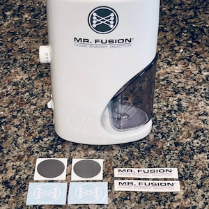 May include: A white "Mr. Fusion Home Energy Reactor" with a black and white logo and text. The device has a clear, angled compartment and a dial with numbers. Several stickers with the logo and circular designs are below the device.