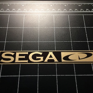 May include: A gold and black sticker with the text "SEGA CD" on a black background.