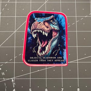 May include: A rectangular sticker with a red border features a detailed illustration of a Tyrannosaurus Rex head with an open mouth. The text "OBJECTS IN MIRROR ARE CLOSER THAN THEY APPEAR" is below the dinosaur image.