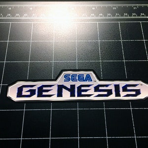 Könnte beinhalten: Ein silbernes und blaues Sega Genesis Logo mit dem Text "SEGA" in Blau und "GENESIS" in Schwarz.