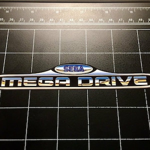 Può includere: Un logo Sega Mega Drive argento e oro con un logo Sega blu e bianco all'interno di un ovale argentato.