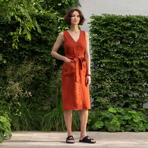 May include: A sleeveless, rust-colored linen dress with a V-neck, button-down front, and a matching tie belt. The dress features two front pockets and is knee-length. The model is wearing black sandals. The background includes lush green foliage.