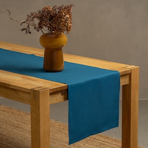 The menique cobalt blue color linen table runner on top of a wooden table. There is a vase on top of the runner.