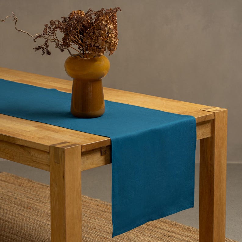 The menique cobalt blue color linen table runner on top of a wooden table. There is a vase on top of the runner.