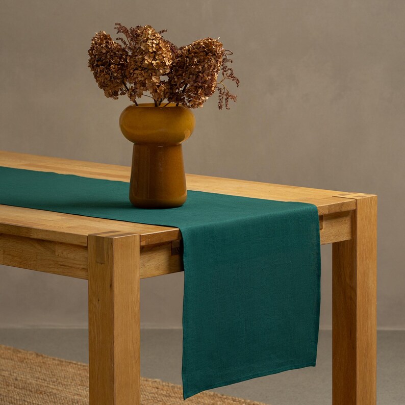 The menique dark green color linen table runner on top of a wooden table. There is a vase on top of the runner.