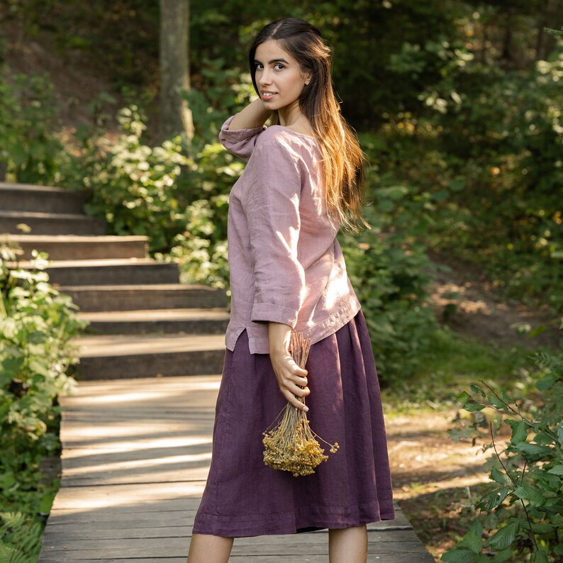 The model is wearing the SOPHIA skirt in shadow purple. She has her back facing the camera, holding a bouquet of wildflowers in her hand by her side. She is facing the camera, wearing a faded rose color long sleeve blouse. The skirt length is midi.