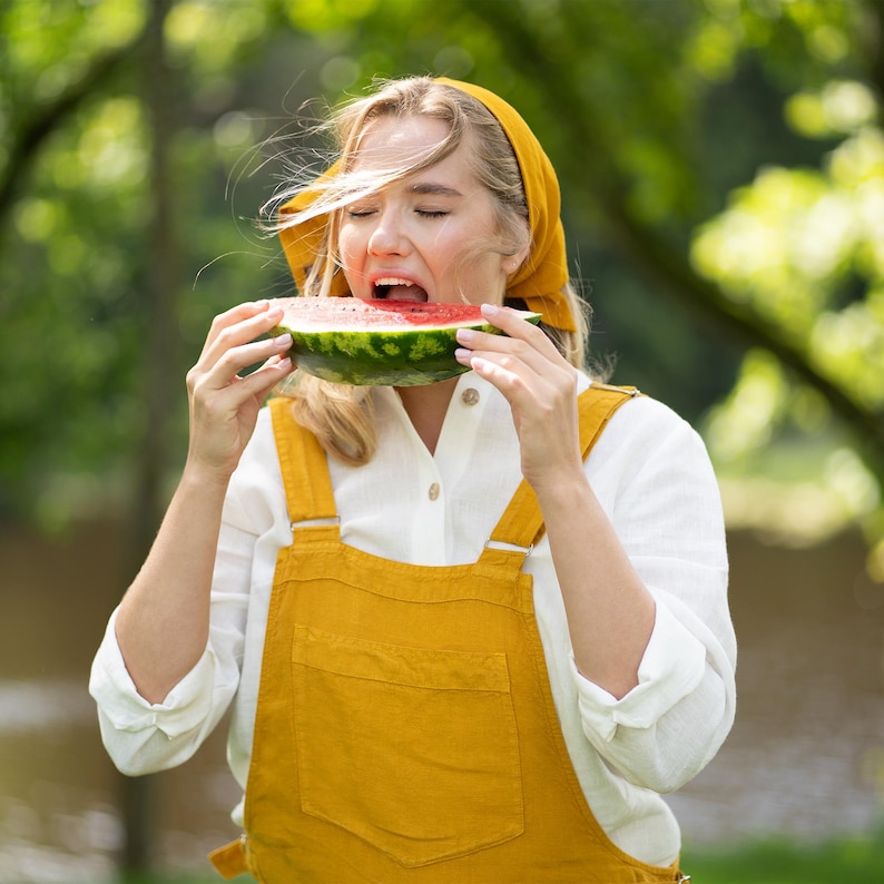 A woman stands outdoors with her eyes closed as she takes a joyful bite out of a large slice of fresh watermelon. She is wearing a spicy yellow linen jumpsuit paired with a crisp white shirt and a matching yellow bandana.