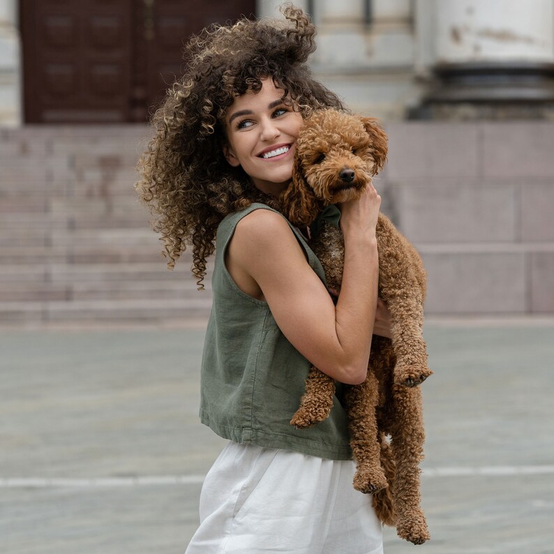 The model is wearing a linen clothing set, which includes a sleeveless crop top in stone green color and white high-waisted pants with pockets. She is holding a dog, smiling, looking sideways.