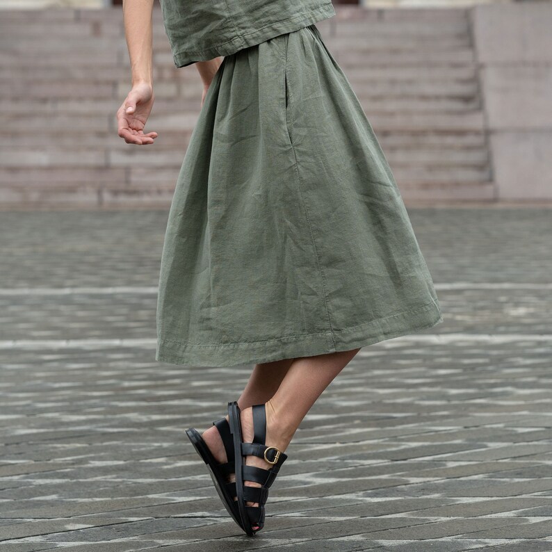 May include: A woman wearing a green linen skirt and black sandals with a gold buckle. The skirt has a pleated design and a side pocket.