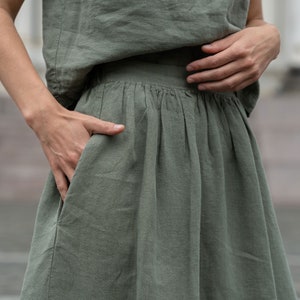The model is wearing the SOPHIA skirt in stone green. This is a close-up picture of the skirt, featuring the side pocket. The woman has her hand inside it.