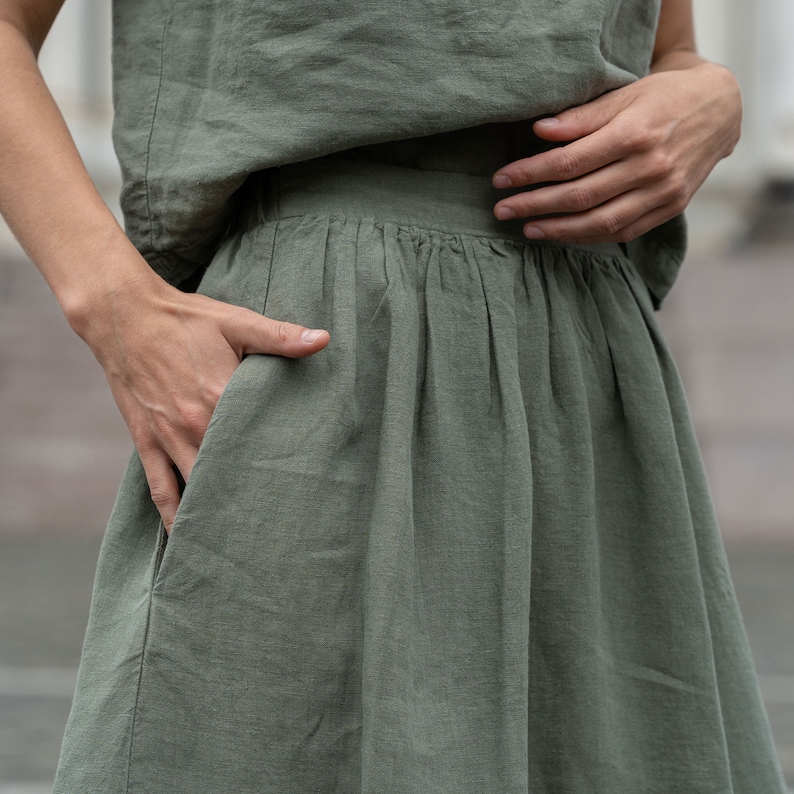 The model is wearing the SOPHIA skirt in stone green. This is a close-up picture of the skirt, featuring the side pocket. The woman has her hand inside it.