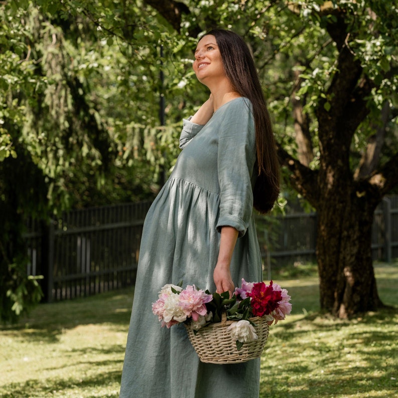The model is wearing the LUCY smock dress in mint green color. The picture shows a pregnant woman in a park. She is standing sideways, the expecting belly highlights the roomy design of the skirt.