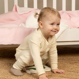 May include: A long-sleeved, cream-coloured shirt and matching trousers set worn by a toddler. The outfit features a simple crew neckline, ribbed cuffs at the wrists and ankles, and a plain, solid-coloured design made of soft, lightweight fabric.