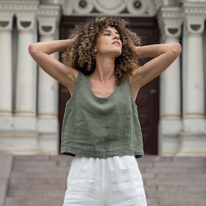 The model is wearing a linen clothing set, which includes a sleeveless crop top in stone green color and white high-waisted pants with pockets. She has both of her hands on the sides of her head.