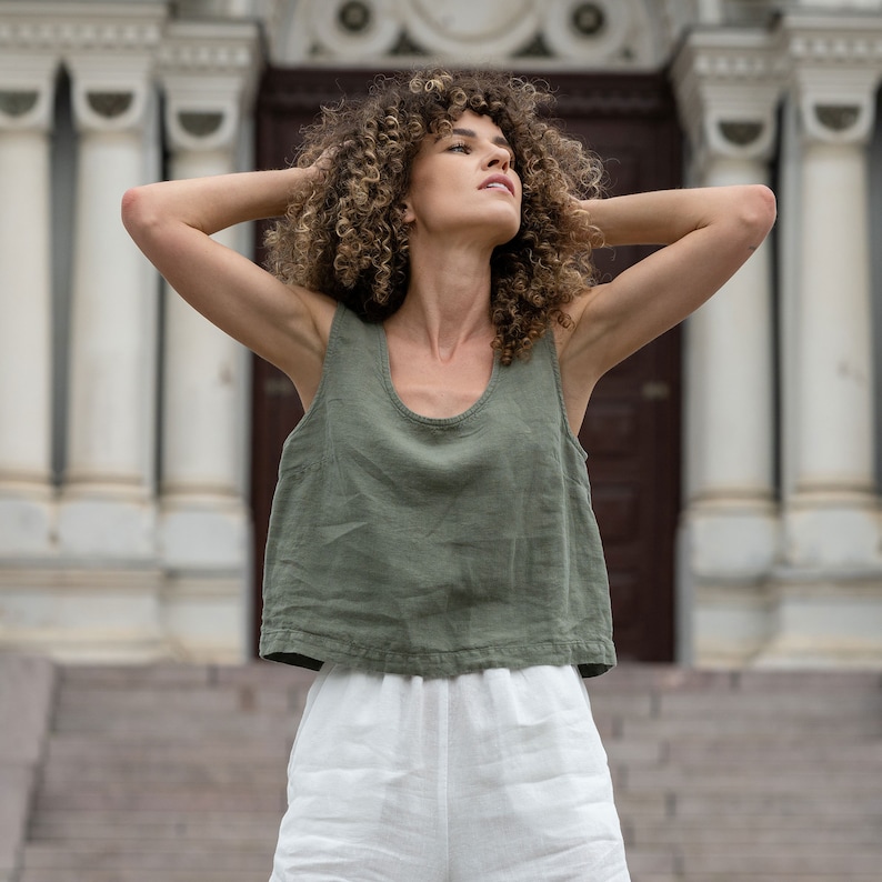 The model is wearing a linen clothing set, which includes a sleeveless crop top in stone green color and white high-waisted pants with pockets. She has both of her hands on the sides of her head.