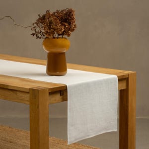 The menique pure white color linen table runner on top of a wooden table. There is a vase on top of the runner.